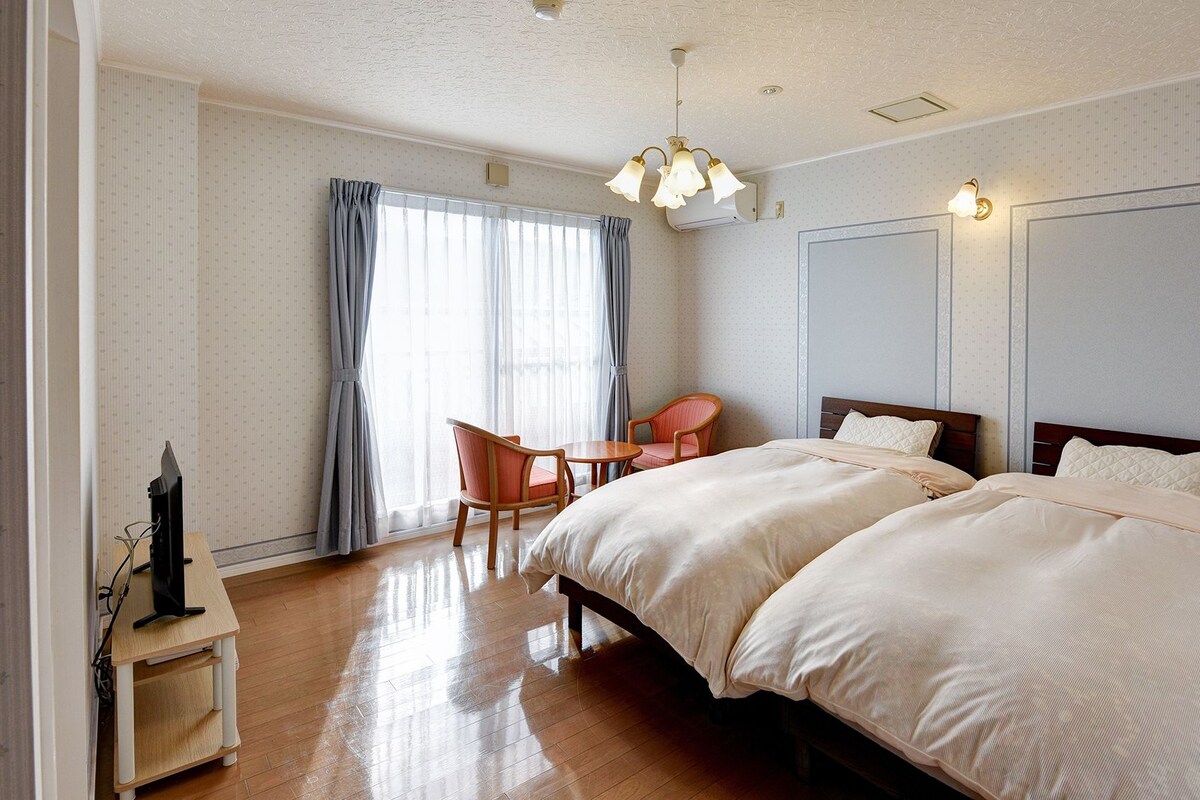 Habitación de hotel con dos camas, un escritorio con silla, una ventana con cortinas y una lámpara en el techo.
