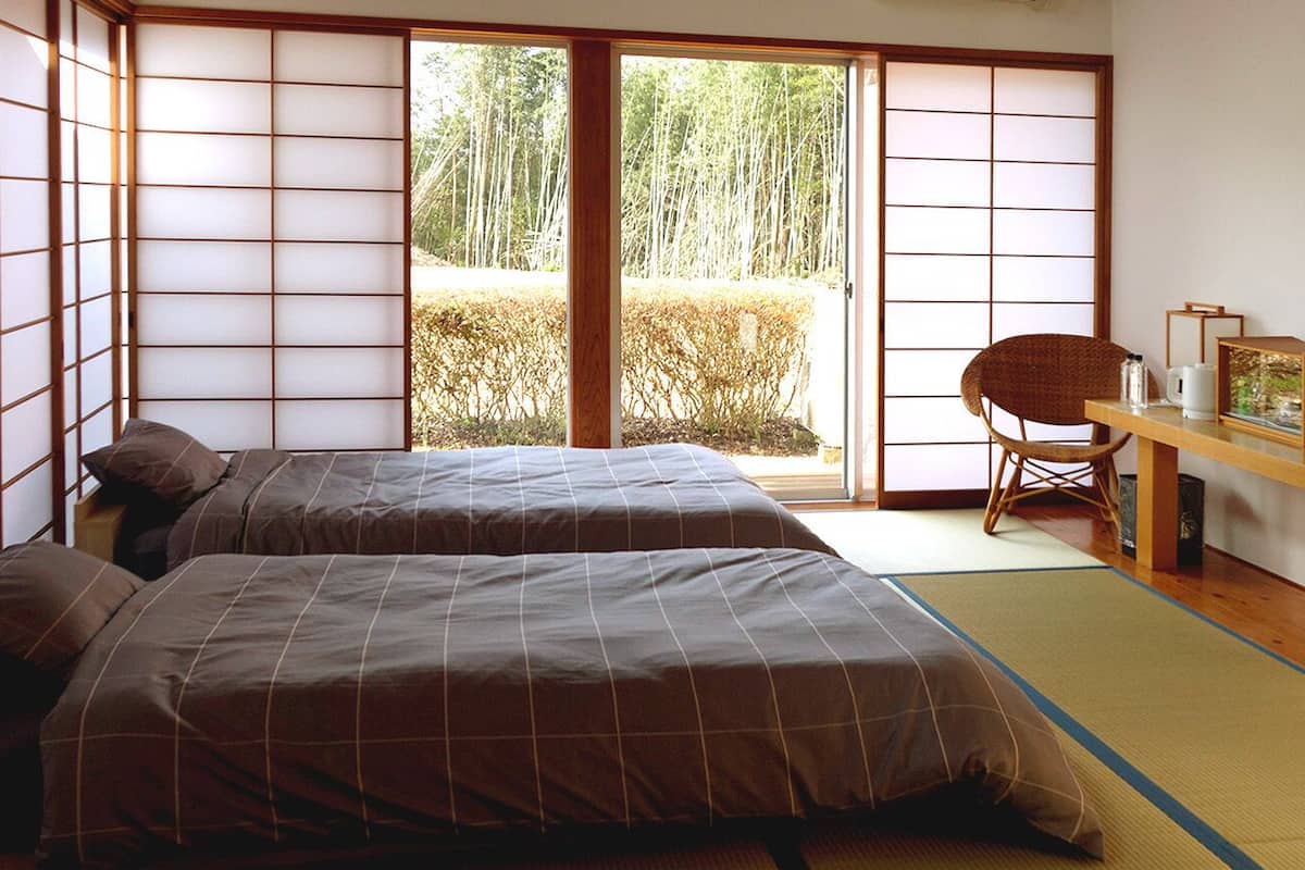 A traditional Japanese room with tatami flooring, a sliding door leading to a garden, two beds with brown bedding, a wooden desk with a television, and a woven chair.
