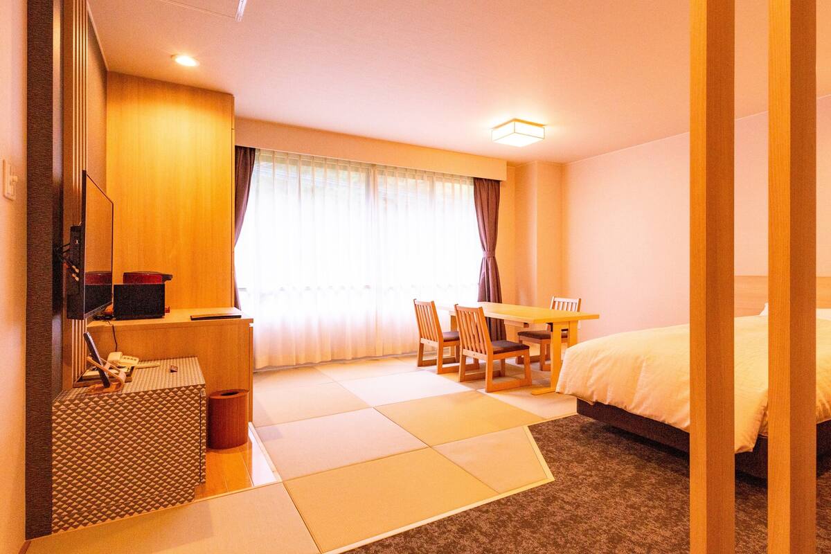A hotel room with a bed, a desk with a chair, a television, and a window with curtains.