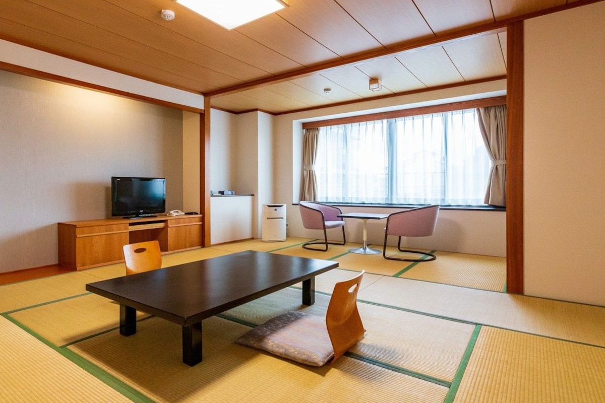 Ein traditioneller japanischer Raum mit Tatami-Boden, einem niedrigen Tisch, einem Fernseher und einem Fenster mit Vorhängen.