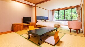A hotel room with a bed, a desk with a television, a chair, and a view of greenery through the window.