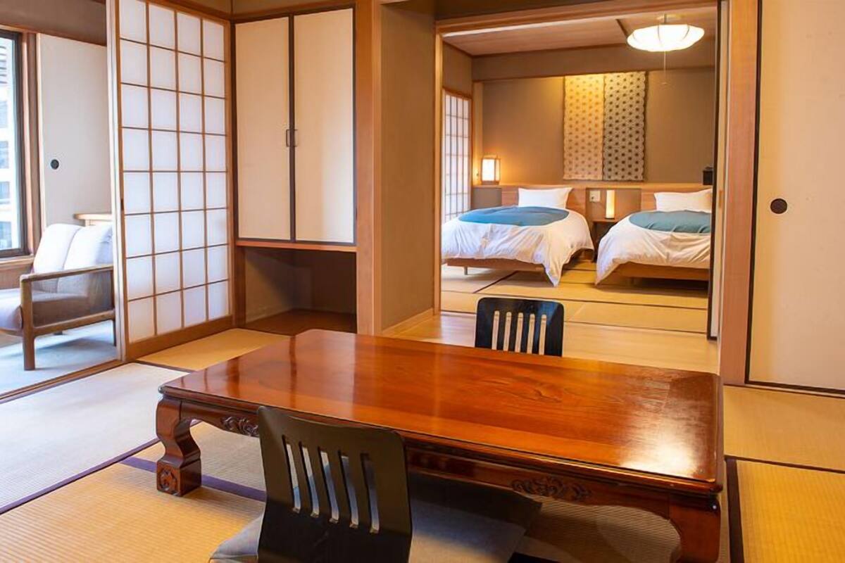 Une pièce de style japonais traditionnel, avec un sol recouvert de tatamis, une petite table en bois, une chaise et une porte coulissante donnant sur une chambre à coucher équipée de deux lits.