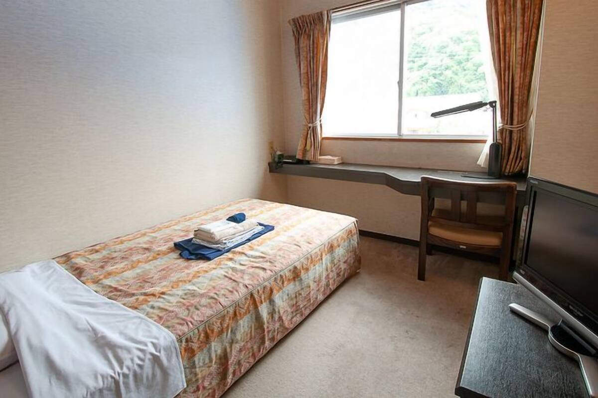 Une petite chambre d’hôtel avec un lit simple, un bureau avec une chaise, une télévision et une fenêtre avec des rideaux.
