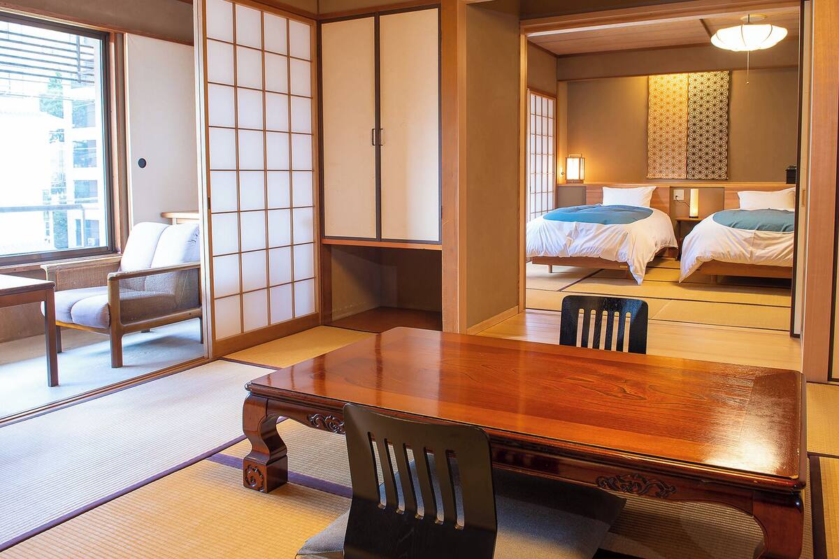 Une pièce de style japonais traditionnel, avec un sol recouvert de tatamis, une table en bois, une chaise, une porte coulissante et un lit doté d’une tête de lit à motifs.