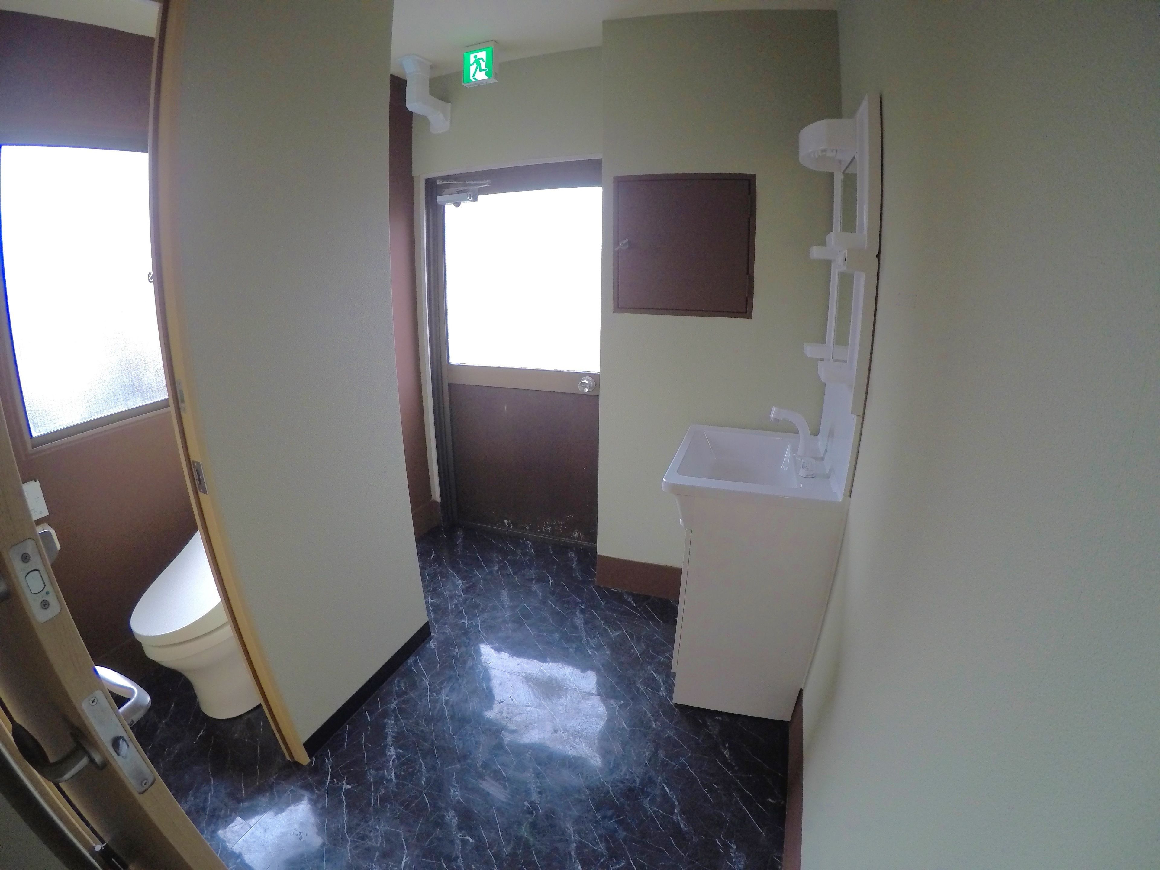 Un baño con inodoro, lavamanos y espejo. Hay una puerta que da a otra habitación y una ventana que deja entrar luz natural.