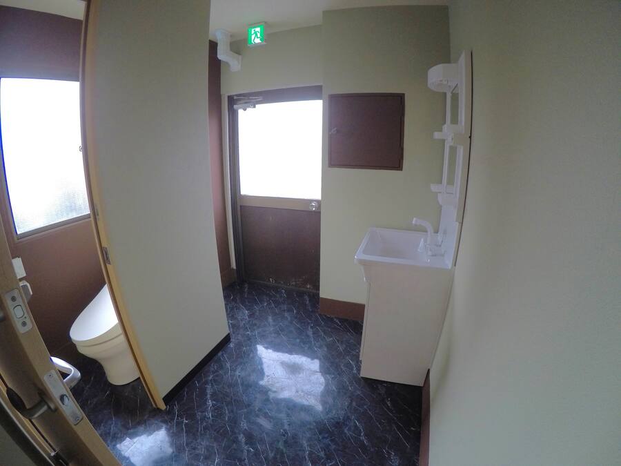 Un baño con inodoro, lavamanos y espejo. Hay una puerta que da a otra habitación y una ventana que deja entrar luz natural.