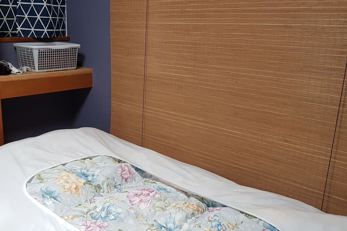 Un dormitorio con estructura de cama de madera, colchón blanco y colcha estampada con motivos florales. Hay un estante de madera con una cesta de ropa y un objeto de diseño geométrico encima.