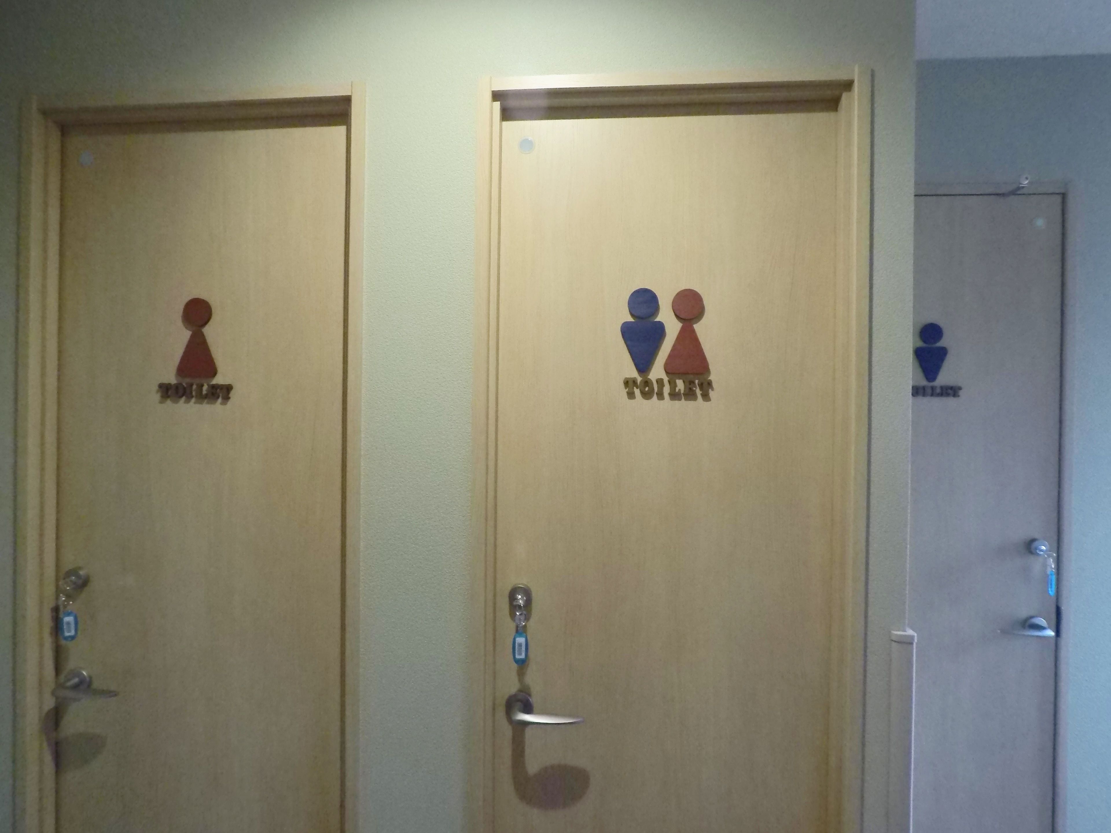 Tre dörrar av trä, försedda med symboler för toaletter, och var och en är markerad med en annan färg för att särskilja toaletterna för respektive kön.