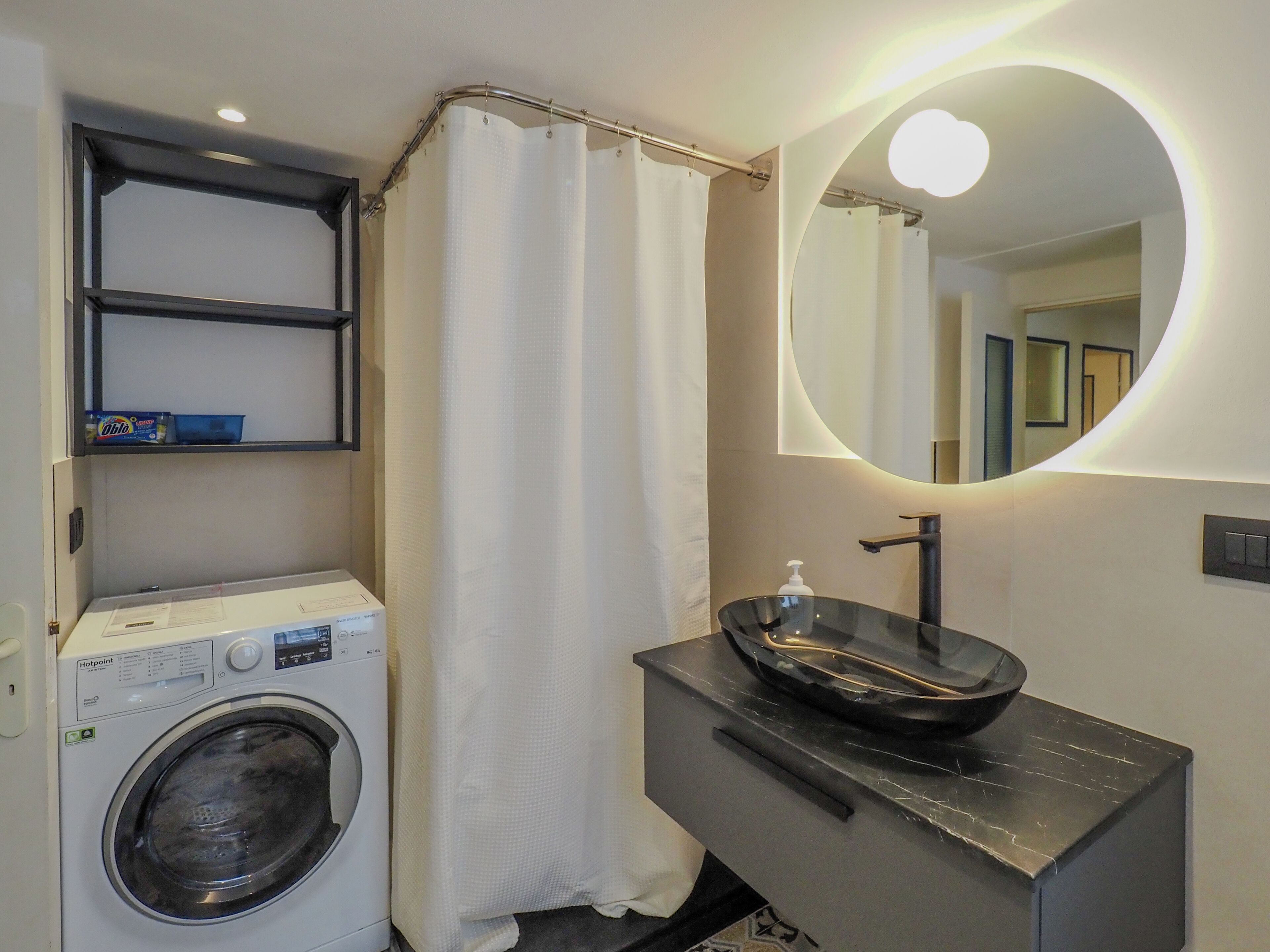 A compact bathroom with a washing machine, a sink with a modern design, and a mirror with a circular light.