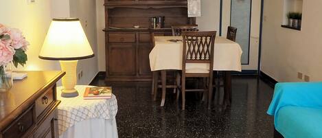 A hotel room with a dining table, chairs, a cabinet with glass doors, a bed, a lamp, and a small table with a book and a vase of flowers.