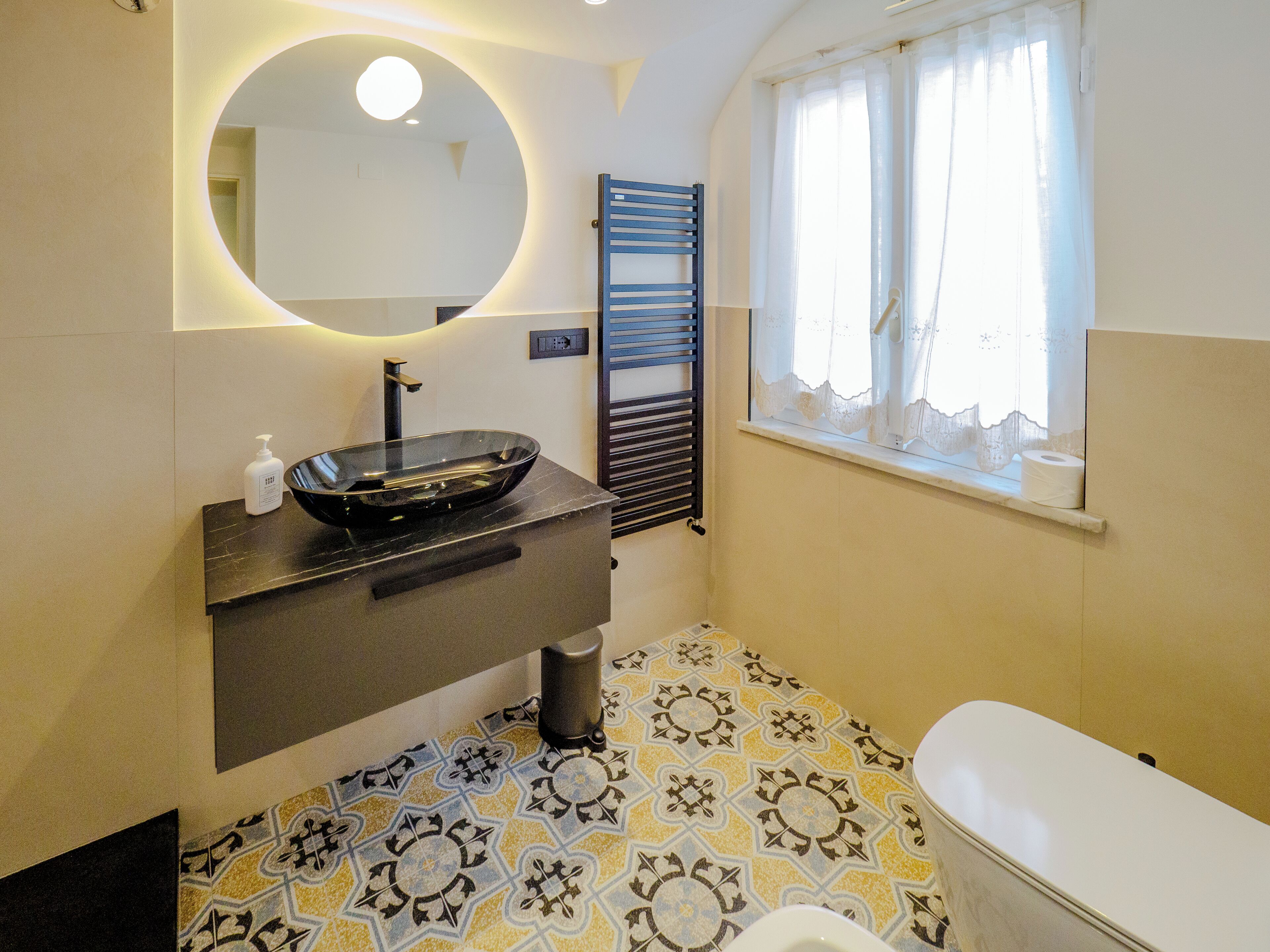 Une salle de bain dotée d’un lavabo moderne, de toilettes, d’une fenêtre avec des rideaux et de carreaux de sol à motifs.