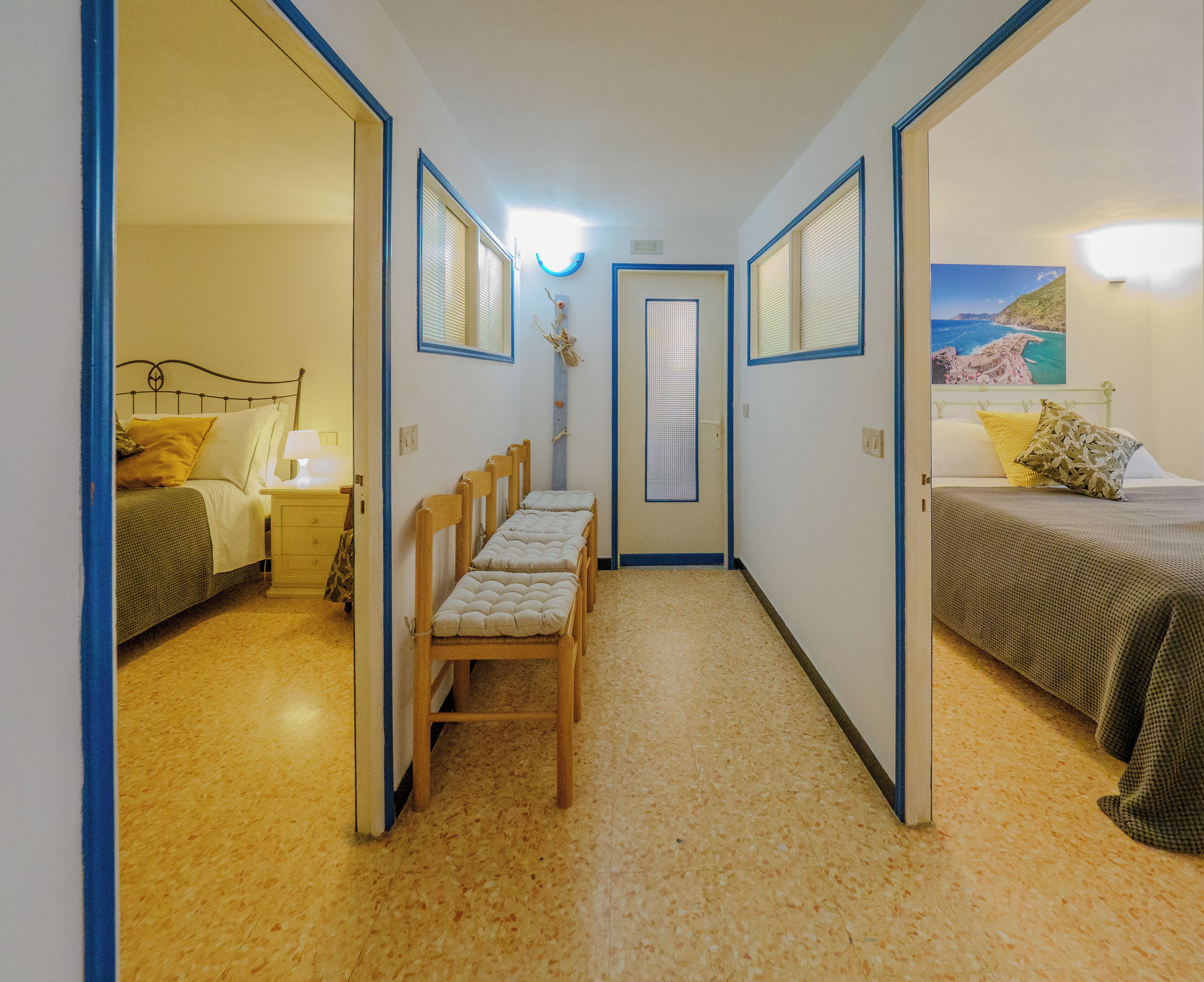 A narrow hallway with a bed on the left and a bed on the right, both with headboards and pillows. There is a small table with chairs in the center of the hallway.