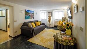 Ein Wohnzimmer mit einer Couch, einem Couchtisch mit Büchern und einer Vase mit Sonnenblumen, einem Bild an der Wand und einem Fenster mit Vorhängen.