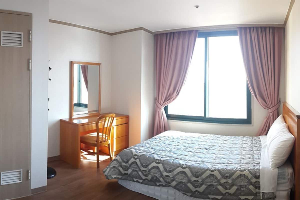 Une chambre à coucher comprenant un lit, un bureau, une chaise, une armoire et une fenêtre avec des rideaux.