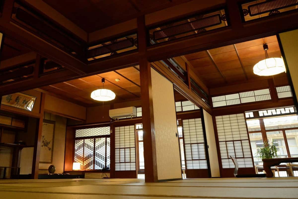 Un intérieur japonais traditionnel avec des poutres en bois, un revêtement de sol en tatami, des portes coulissantes et un luminaire suspendu.