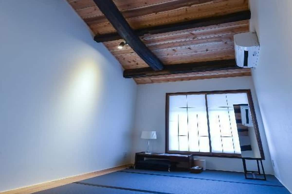 Ein Zimmer mit schrägem Dach, einem Fenster mit Jalousien, einer blauen Matte auf dem Boden und einem kleinen Tisch mit einer Lampe.
