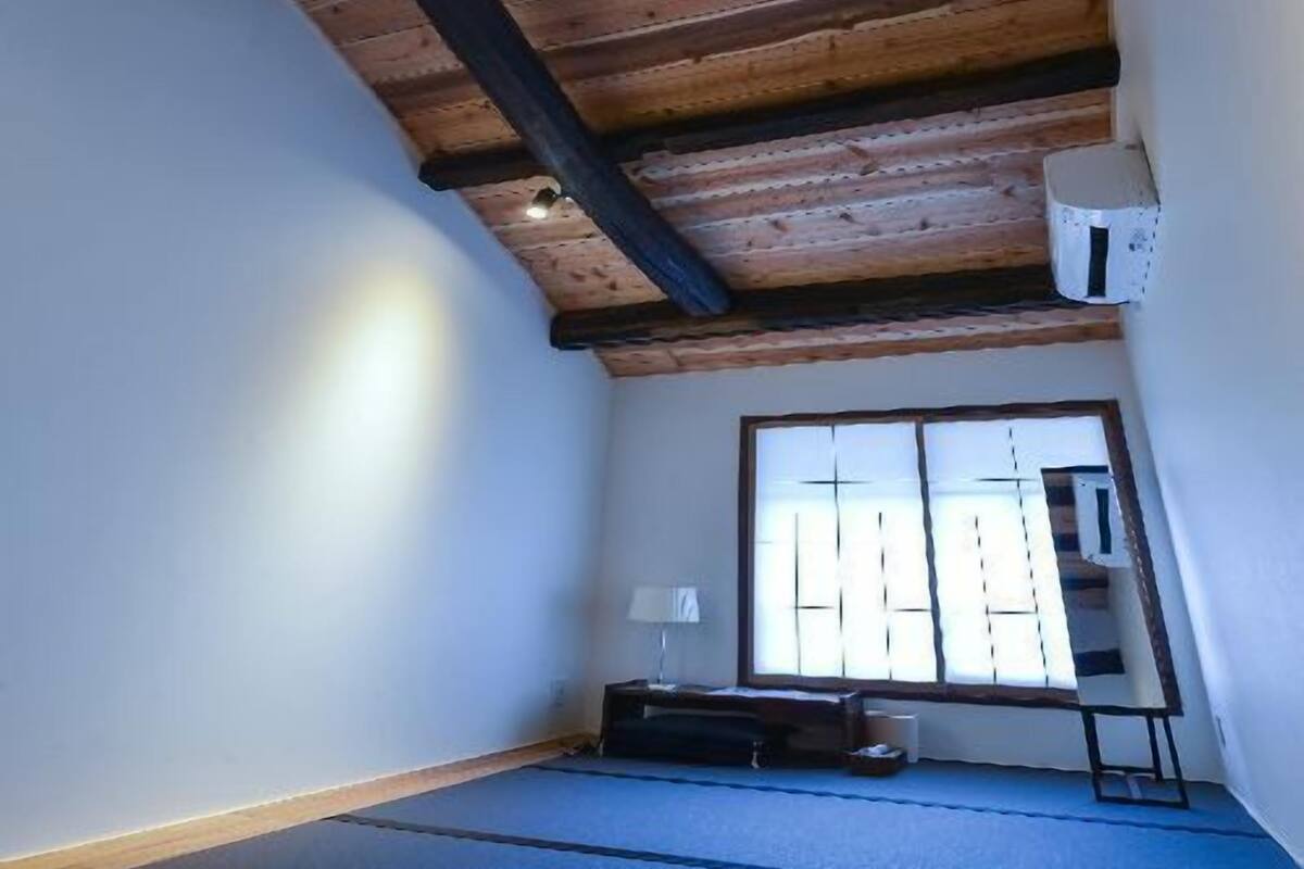 Une pièce avec un plafond en pente, une fenêtre avec des stores, un tapis bleu sur le sol et une petite table avec une lampe.