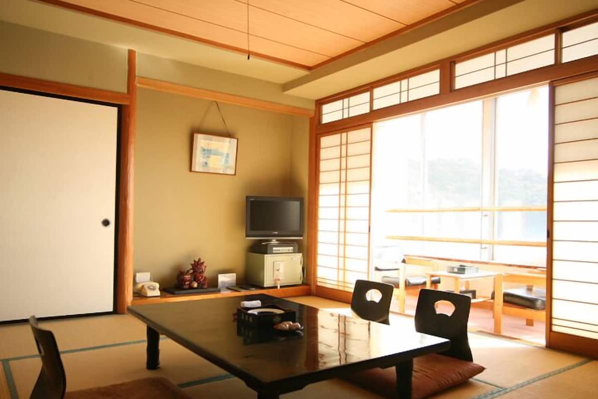 Ein traditioneller japanischer Raum mit Tatami-Boden, einem niedrigen Tisch, Stühlen, einem Fernseher und einer Schiebetür mit Blick.