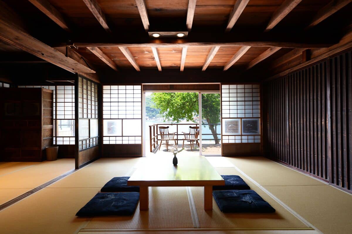 Ein traditioneller japanischer Raum mit Tatami-Böden, einem niedrigen Holztisch und Schiebetüren, die zu einem Außenbereich mit Blick auf Bäume und einem für zwei Personen gedeckten Tisch führen.