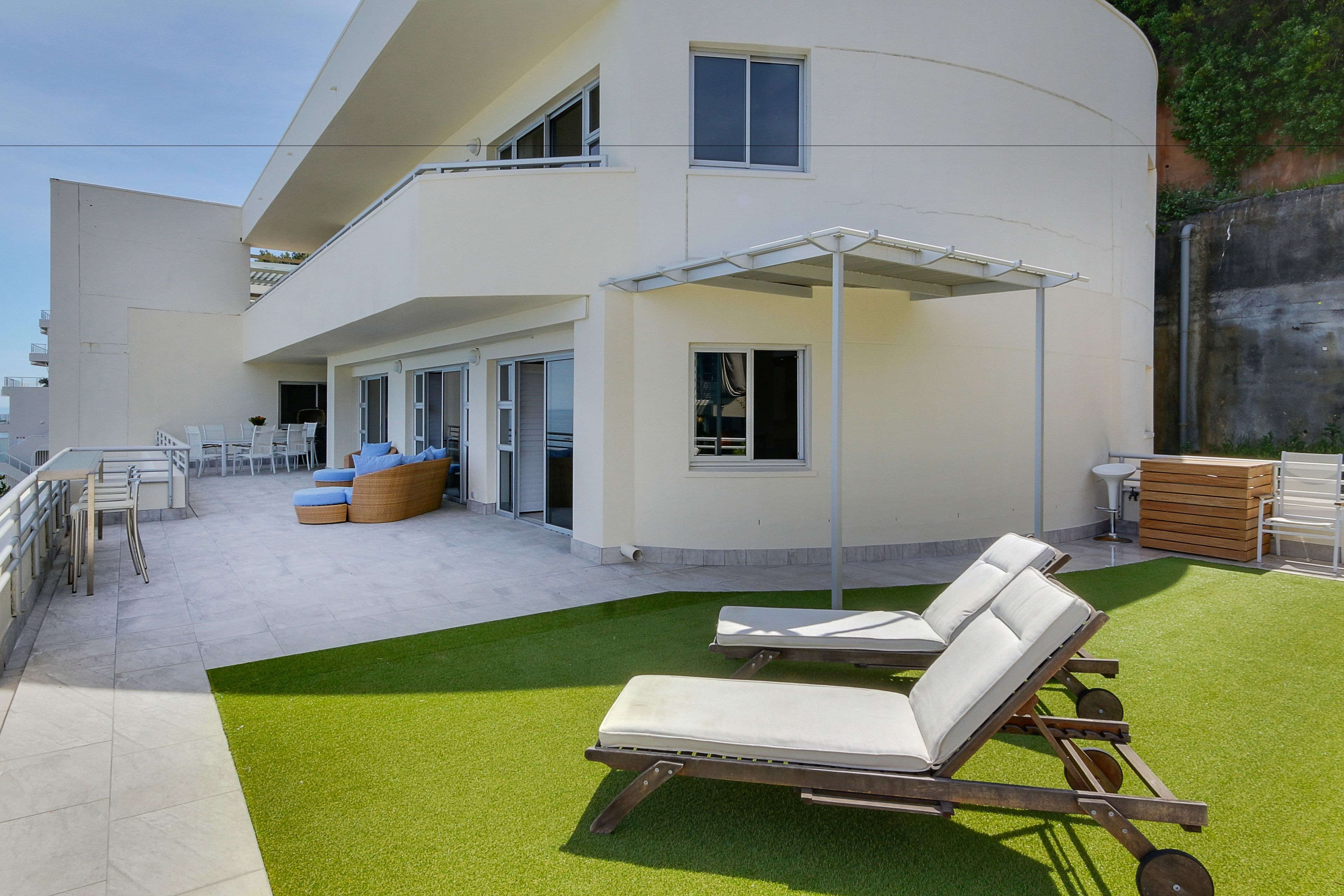 Un espace extérieur moderne comprenant deux fauteuils de relaxation, un coin salon abrité et une terrasse avec, en arrière-plan, un bâtiment blanc.