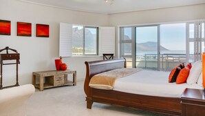 A bedroom with a large bed, a wooden nightstand, a chair, and a view of the ocean through large windows. - Dunmore Views (Cape Town)