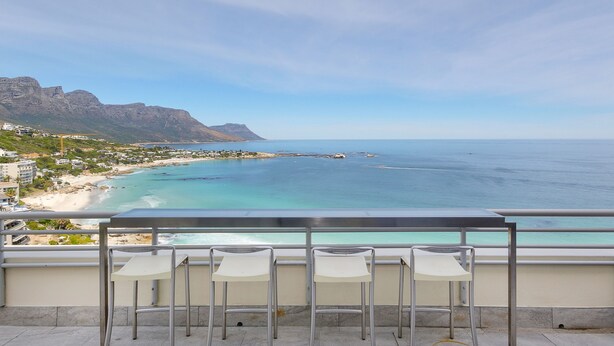 A balcony with four white chairs overlooking a beach with clear turquoise waters and distant mountains. - Dunmore Views (Cape Town)