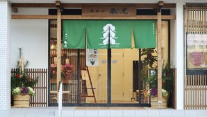A hotel entrance with a green awning displaying the hotel's name and logo. There are potted plants and a wooden fence on either side of the entrance. - Hotel Matsuo (Hamada)