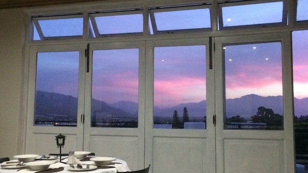 A dining area with a table set for two, large windows with a view of mountains and a sunset, and a wooden ceiling. - Paarl Mountain Lodge (Paarl)