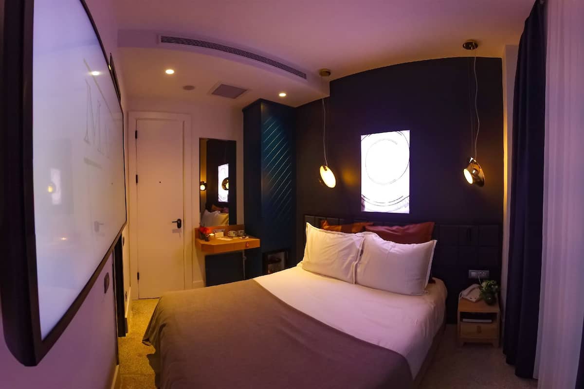 A hotel room with a bed, a bedside table, a nightstand, a mirror, and a window with a view.