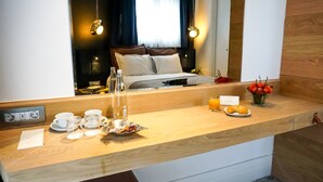 Une chambre d’hôtel avec un comptoir en bois où le petit-déjeuner est servi, un lit avec une tête de lit blanche, un vase de fleurs et une vue sur l’intérieur de la pièce, visible à travers un miroir.