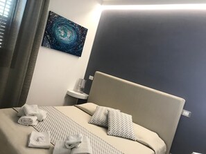 Una cama bien hecha con toallas, una colcha estampada y almohadas, una mesita de noche con lámpara, y un cuadro en la pared.