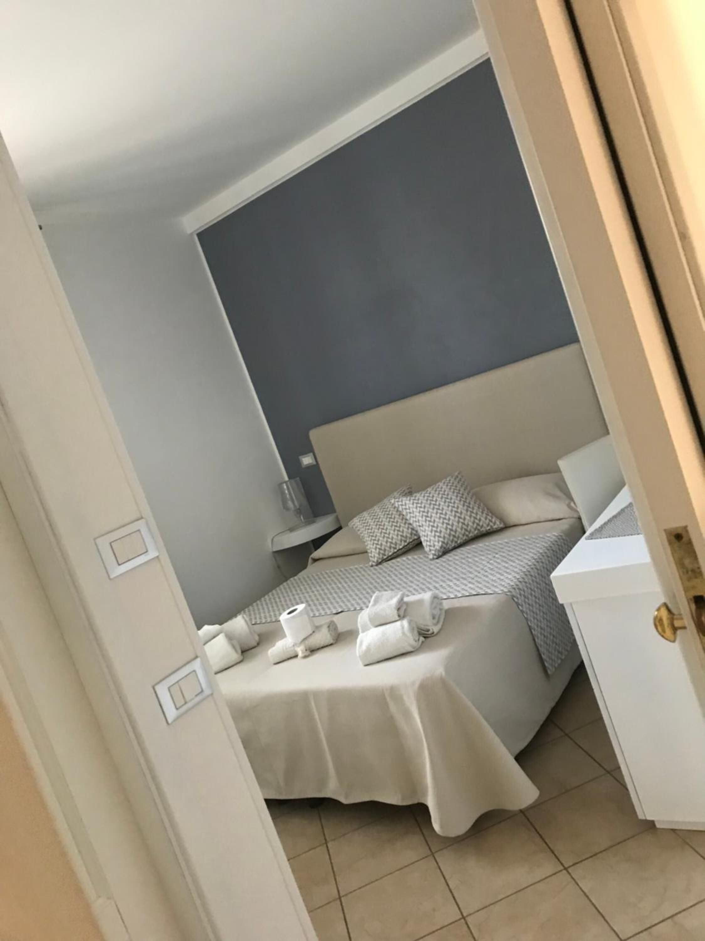 Una cama bien hecha con colcha a cuadros gris y blanca, dos almohadas y toallas enrolladas sobre una mesita de noche blanca.