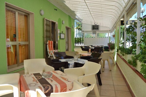 An outdoor cafe with tables and chairs, a covered area with a white ceiling, and a green wall. - Hotel Villa Roberta (Jesolo)