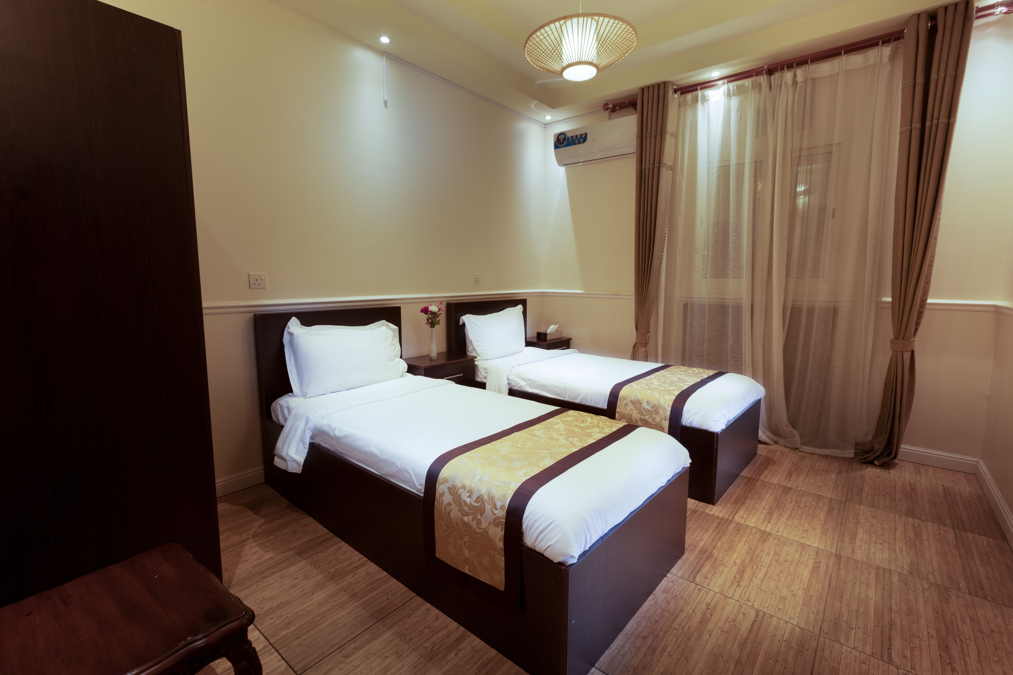 Habitación de hotel con dos camas, suelo de madera, lámpara en el techo y una ventana con cortinas.