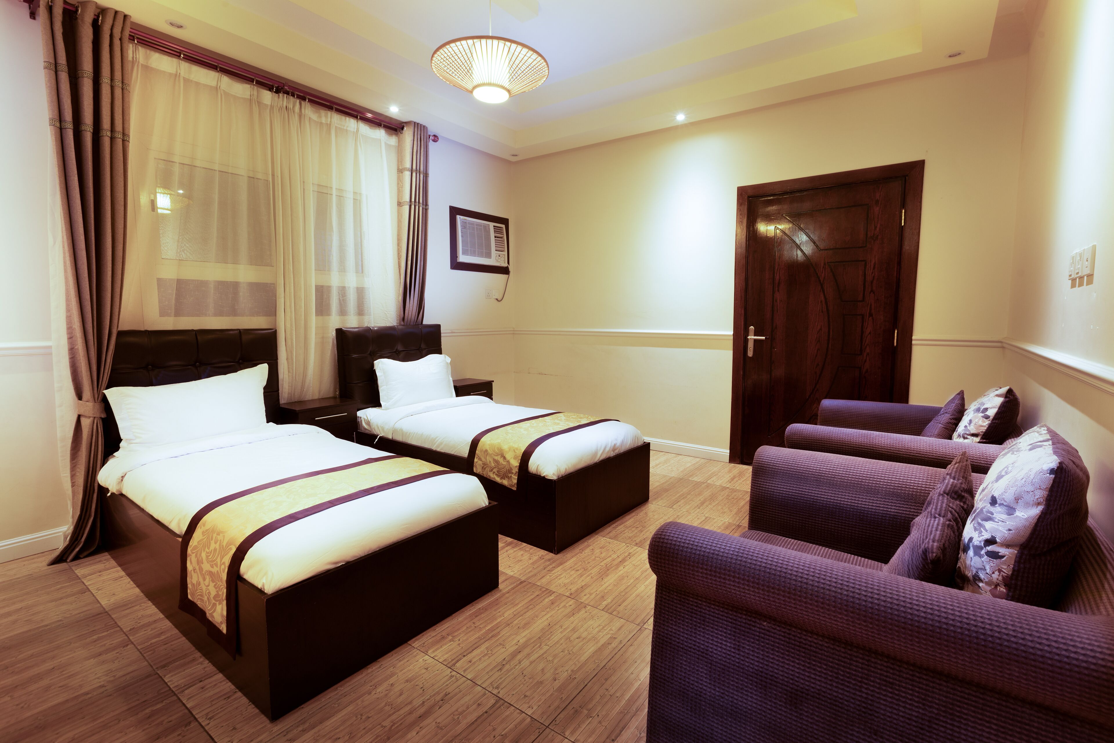 Habitación de hotel con dos camas, un sofá, piso de madera, una lámpara de techo y una puerta.