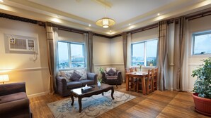 A living room with a sofa, a coffee table, a dining table with chairs, a potted plant, and large windows with curtains. - Gardens View Serviced Apartment  (Taif)