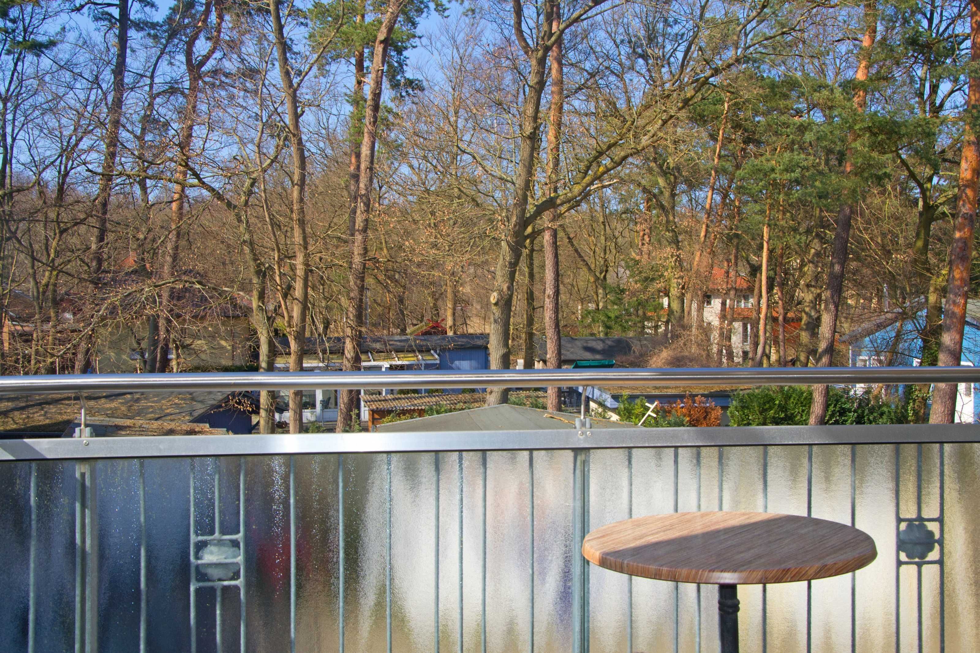 A balcony with a metal railing, a wooden table, and a view of a forested area with trees and a distant building.