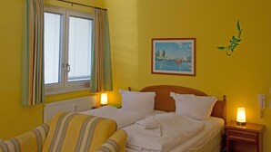 Habitación de hotel con dos camas, un sofá, una ventana con cortinas, un cuadro en la pared y una mesita de noche con lámpara.