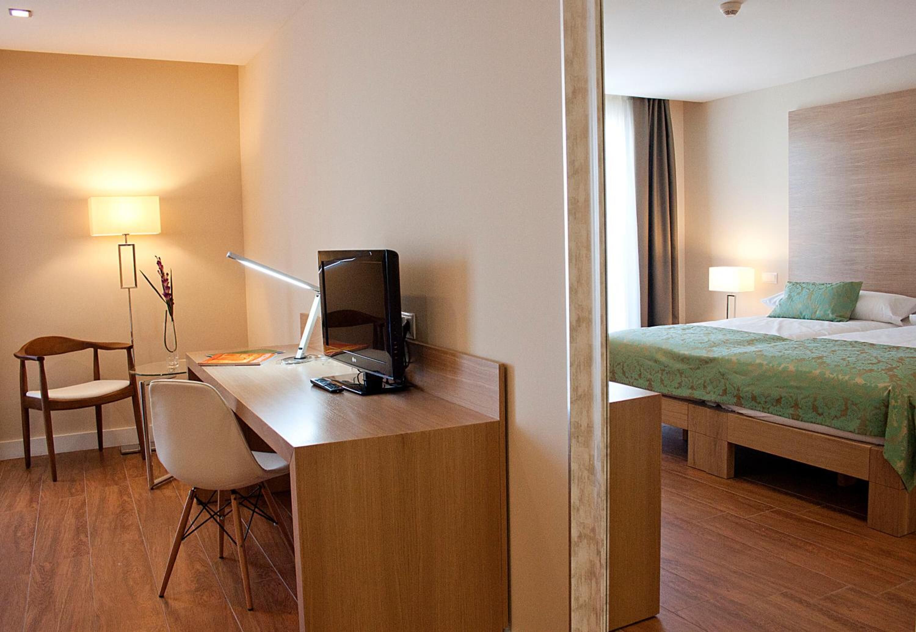 Habitación de hotel con un escritorio de madera, una silla, una cama con ropa de cama verde y una mesita de noche con lámpara.