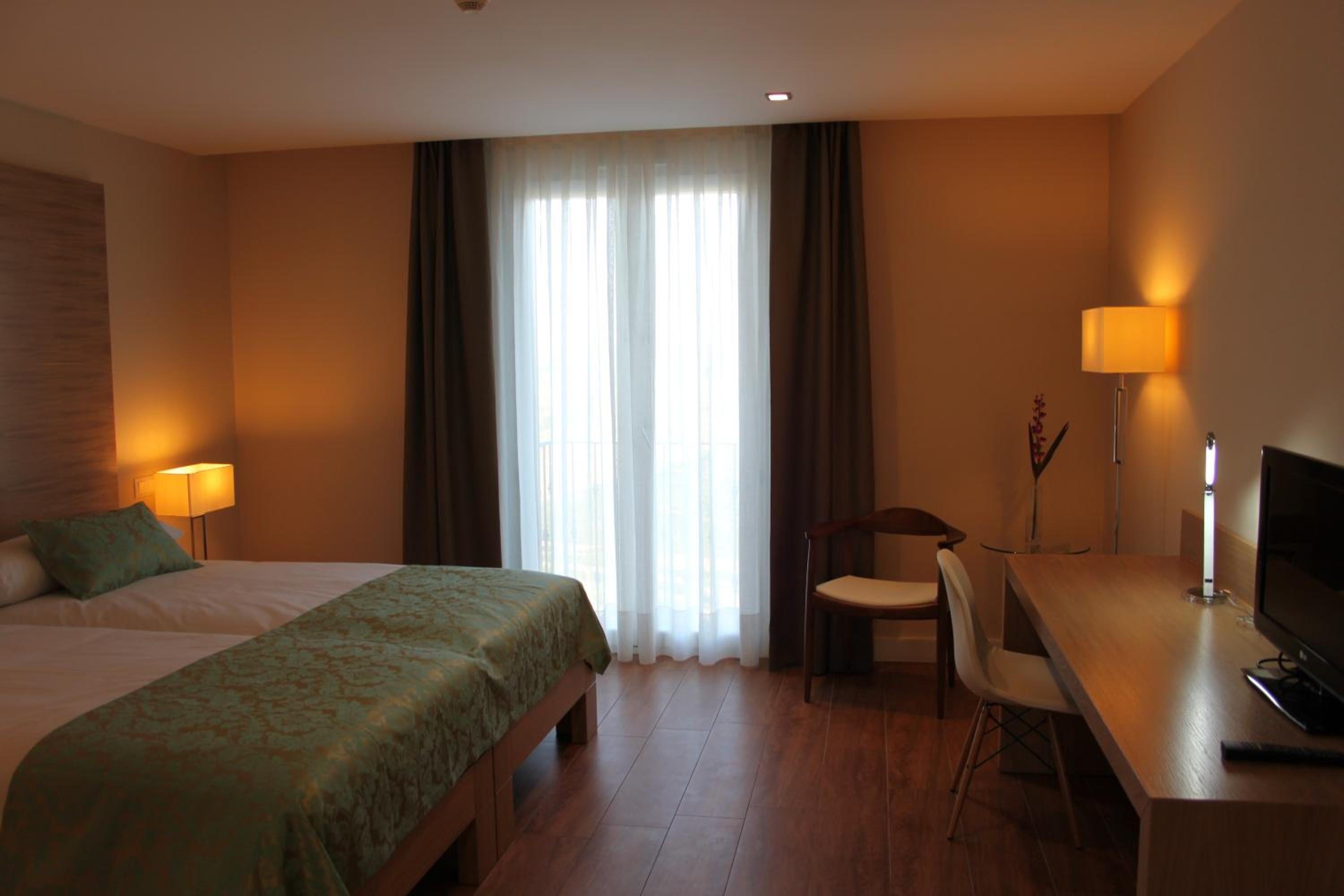 A hotel room with a bed, a desk with a chair, a TV, and a lamp.