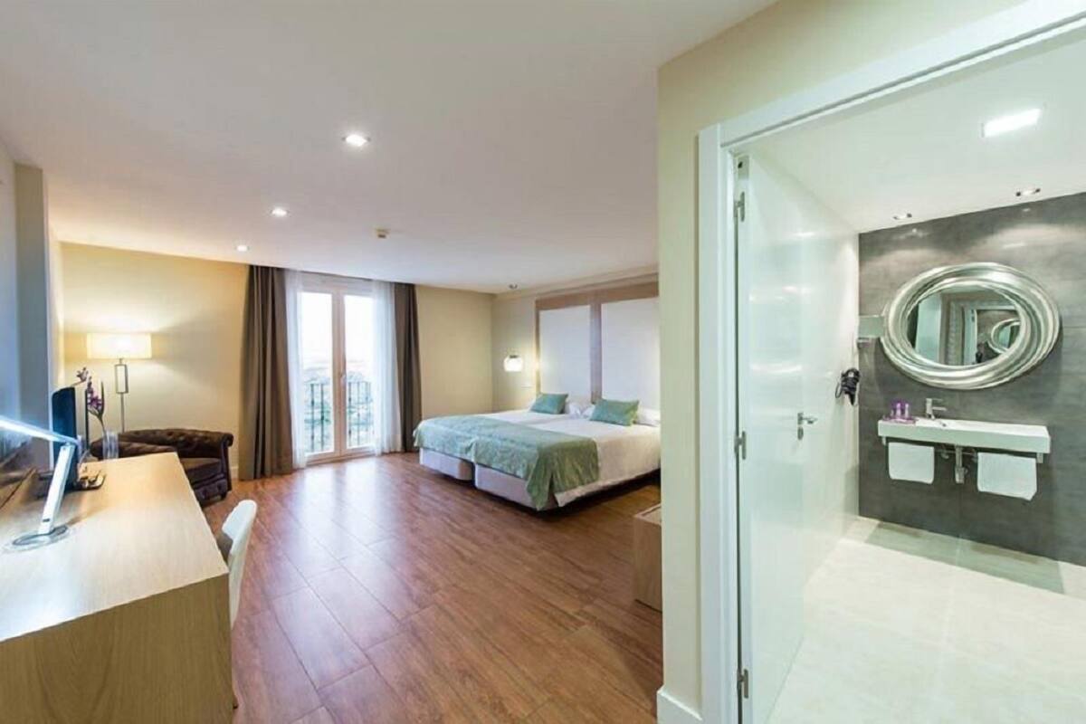 A modern hotel room with a wooden floor, a bed with green bedding, a desk with a lamp, and a bathroom with a mirror and sink.