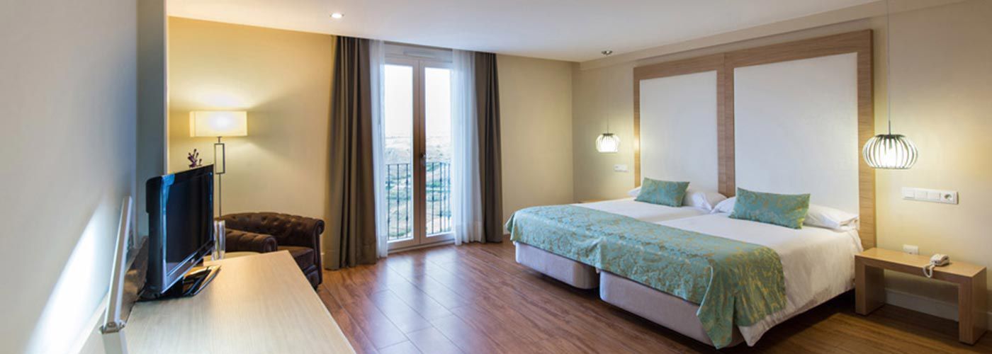 A hotel room with a bed, a desk with a TV, a chair, and a balcony with curtains.