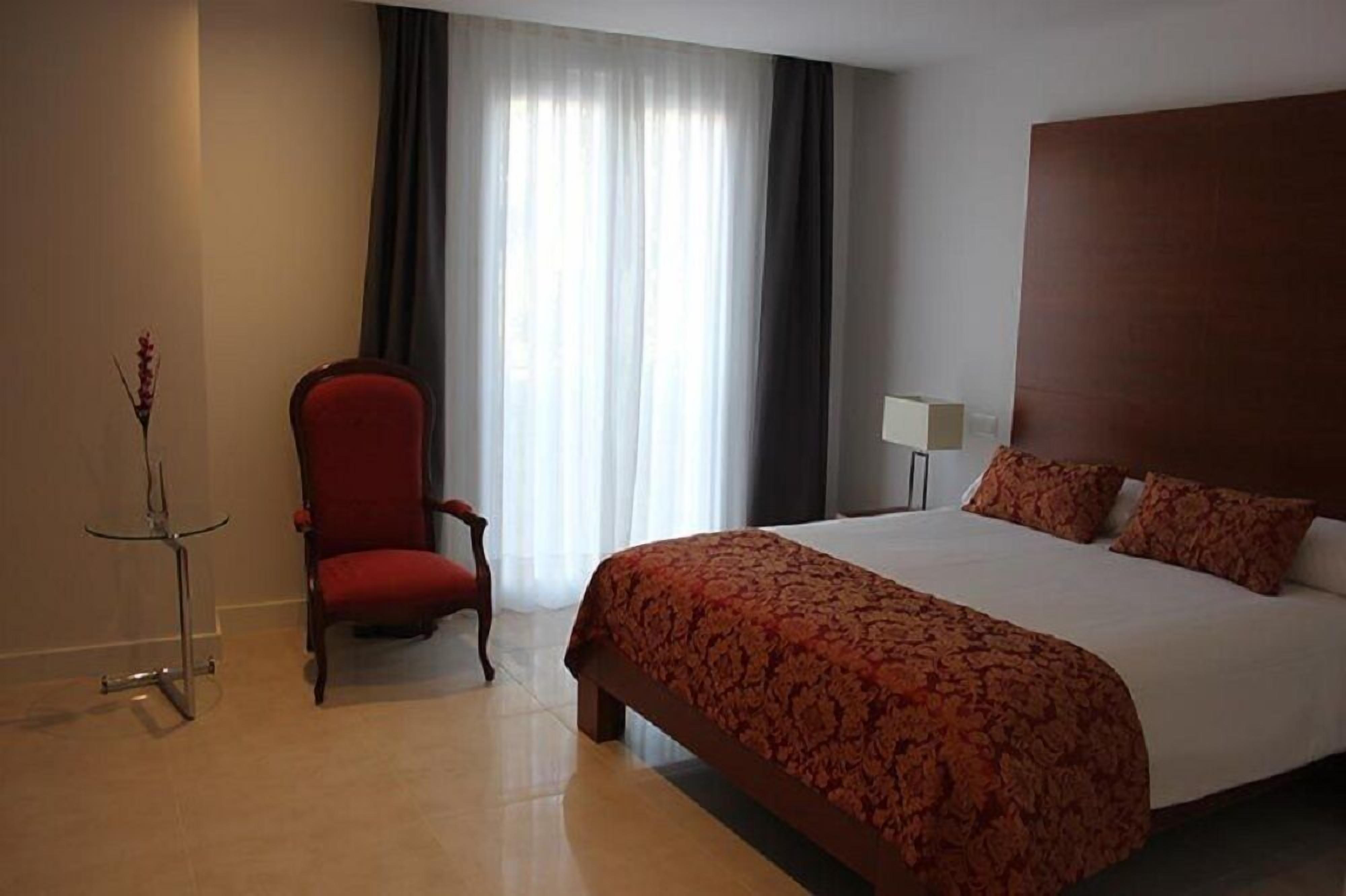 Una habitación de hotel con una cama grande, una silla roja, una mesita pequeña y una ventana con cortinas.