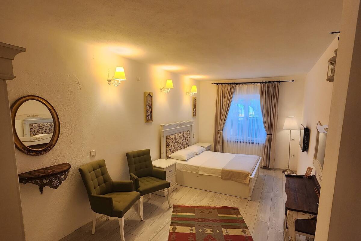 Une chambre à coucher de taille modeste, comprenant un lit, deux chaises, une petite table, un miroir et une fenêtre avec des rideaux.