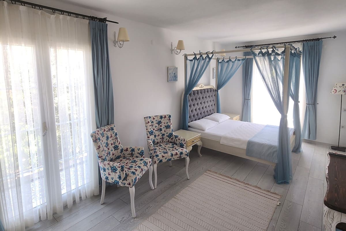 Une chambre à coucher comprenant un lit, deux chaises, une table de chevet, une commode, une lampe et une fenêtre avec des rideaux.