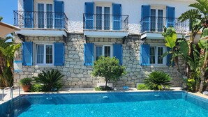 A two-story building with a stone facade and blue shutters, a swimming pool in front, and tropical plants around.