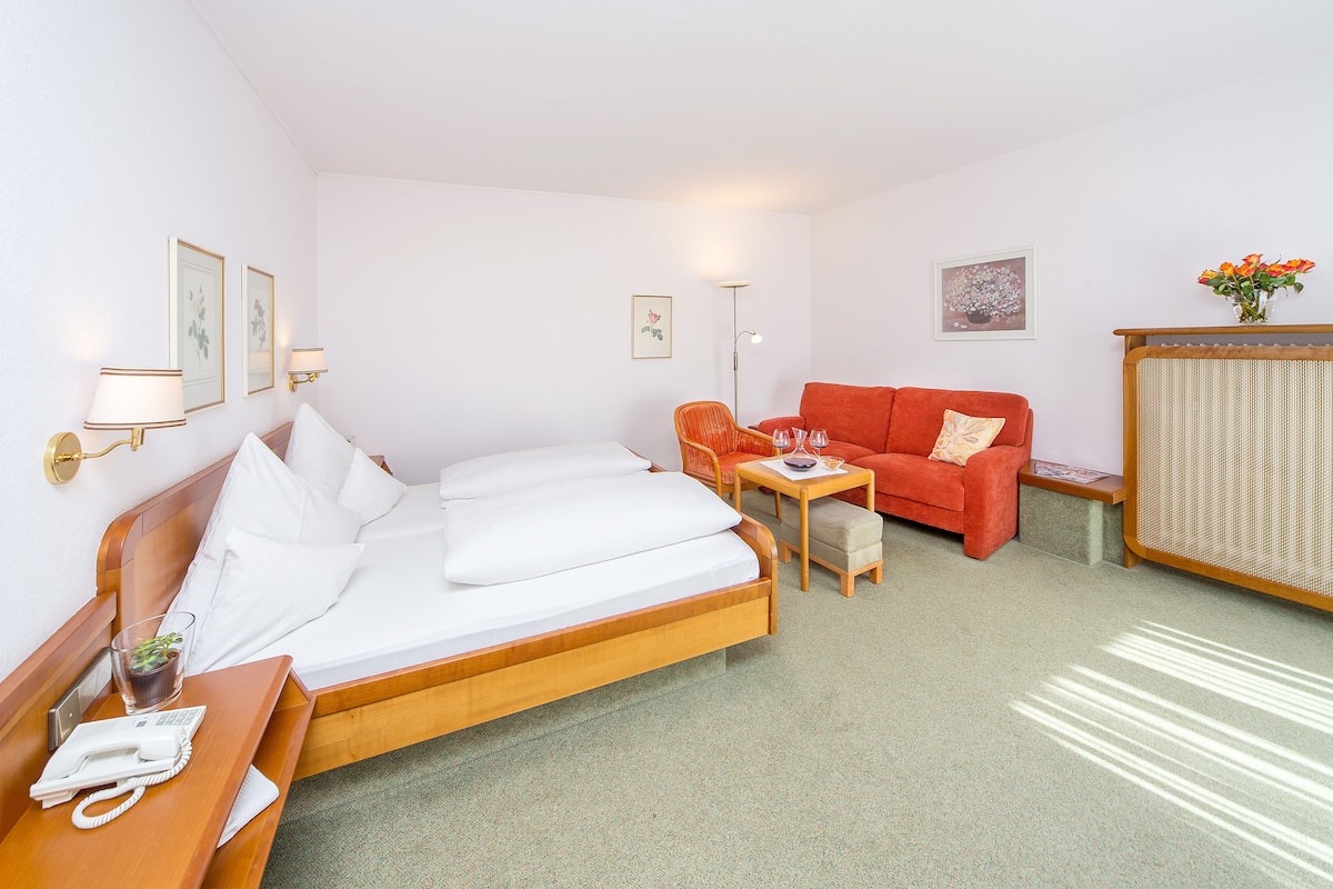 Quarto de hotel com cama, sofá vermelho, mesinha, luminária e um vaso com flores.