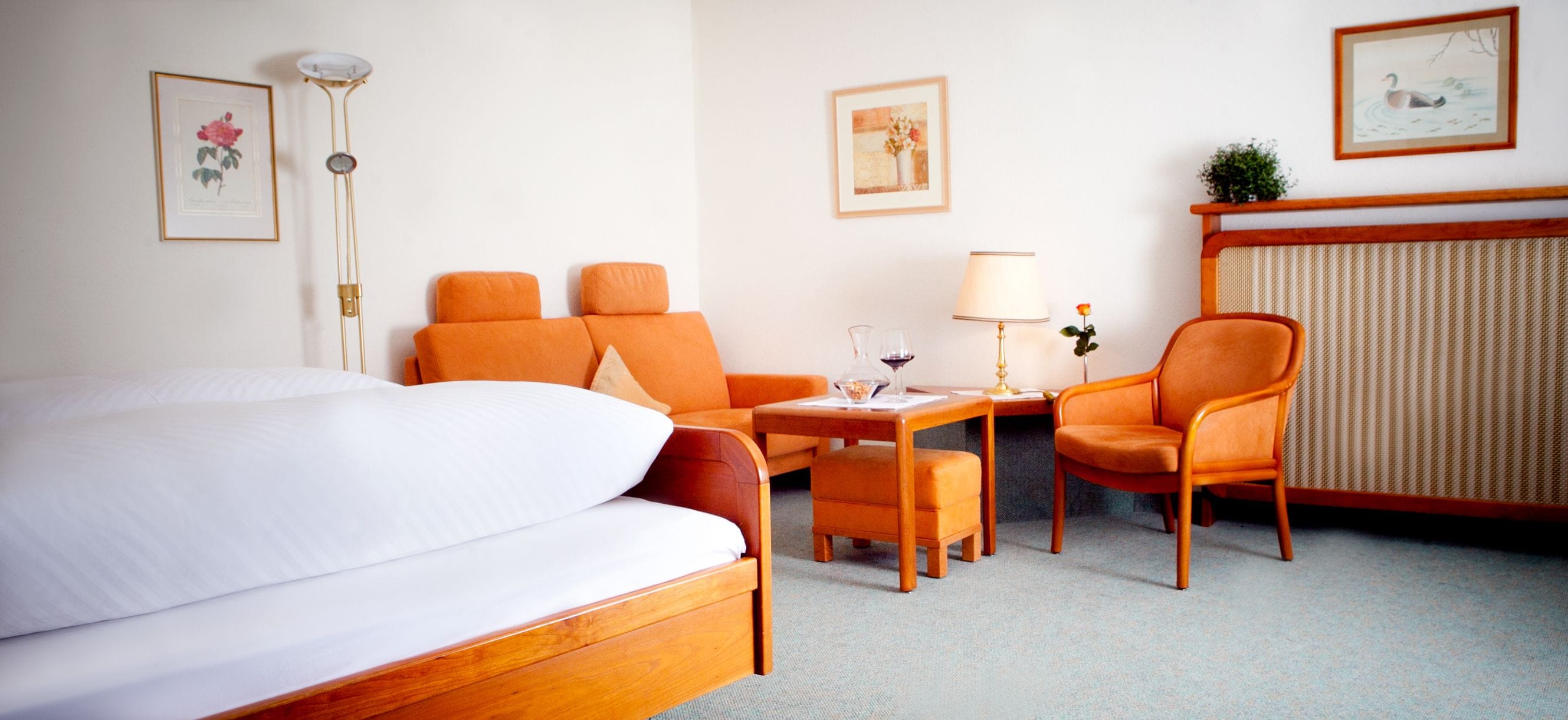 Habitación de hotel con una cama grande, dos sillones naranja, una mesita de noche con lámpara y un radiador.