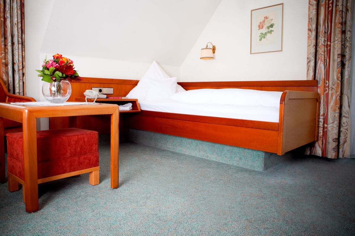 Quarto de hotel com cama de madeira, uma mesinha com um vaso de flores, mesa de cabeceira com abajur e um quadro na parede.