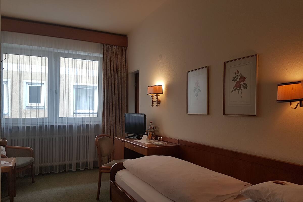 Quarto de hotel com cama, escrivaninha com computador, cadeira e duas luminárias de parede.